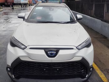 A white compact crossover SUV with a distinctive front grille and sharp LED headlights, parked on a wet pavement near a line of shipping containers. The vehicle features a blue emblem at the center of the grille.