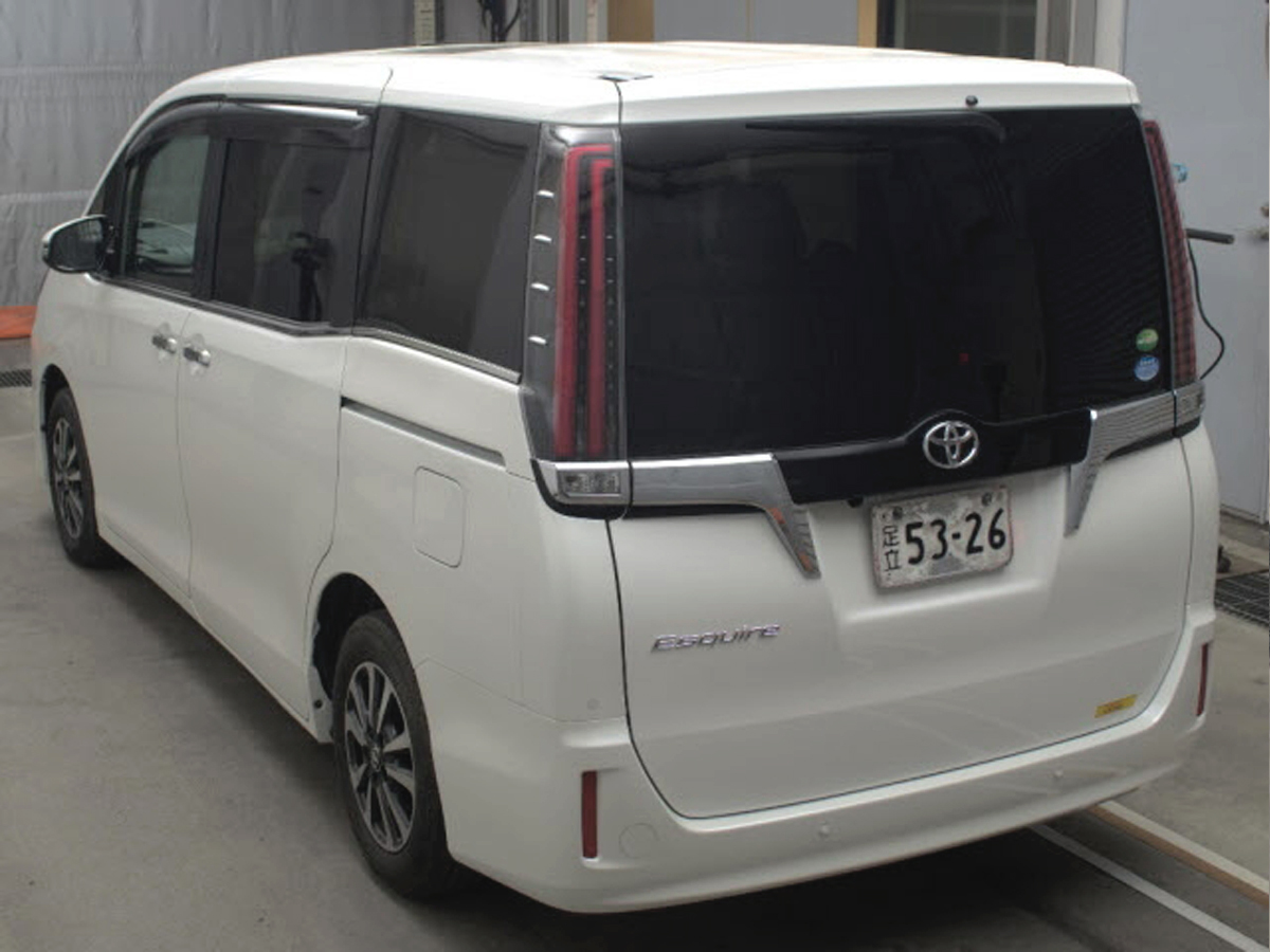Car Planet A luxurious 2019 Toyota Esquire Gi minivan in pristine white is parked indoors. The vehicle features a sleek and modern design, with a prominent chrome grille and sharp, sophisticated headlights. The minivan's clean lines and spacious body are accentuated by large windows and stylish alloy wheels, giving it an elegant and upscale appearance. The license plate and a small "JU" logo are visible, indicating it is from a Japanese auction.